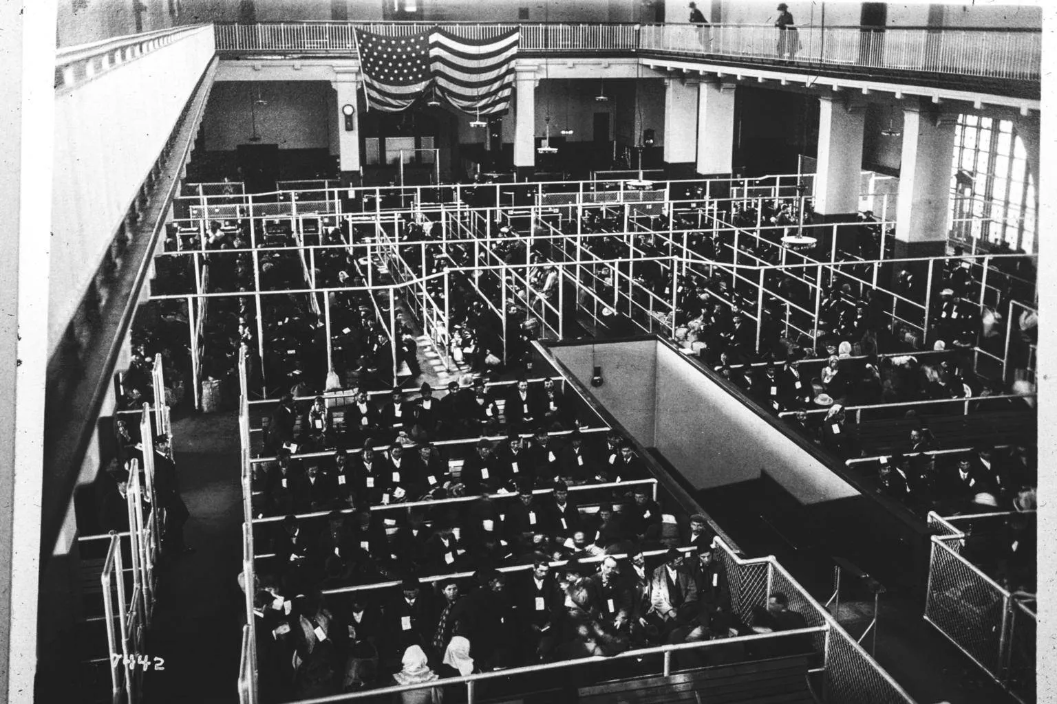 Black-and-white photograph of immigrants in the Ellis Island registry room, standing in pens after the first inspection, about 1902 to 1913.